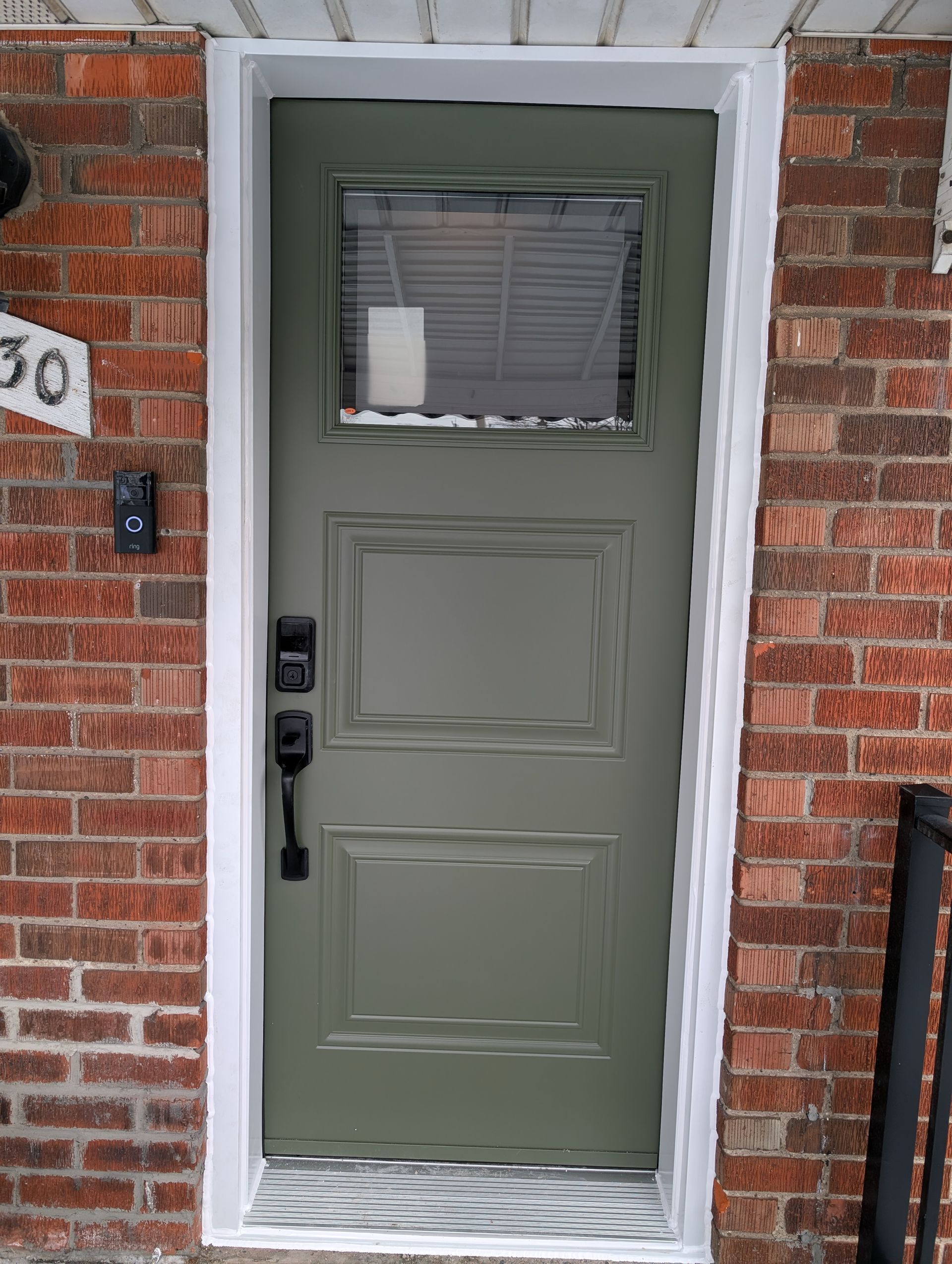 A sage green front door with a glass window at the top, two rectangular panels, and a smart lock, set in a brick wall.