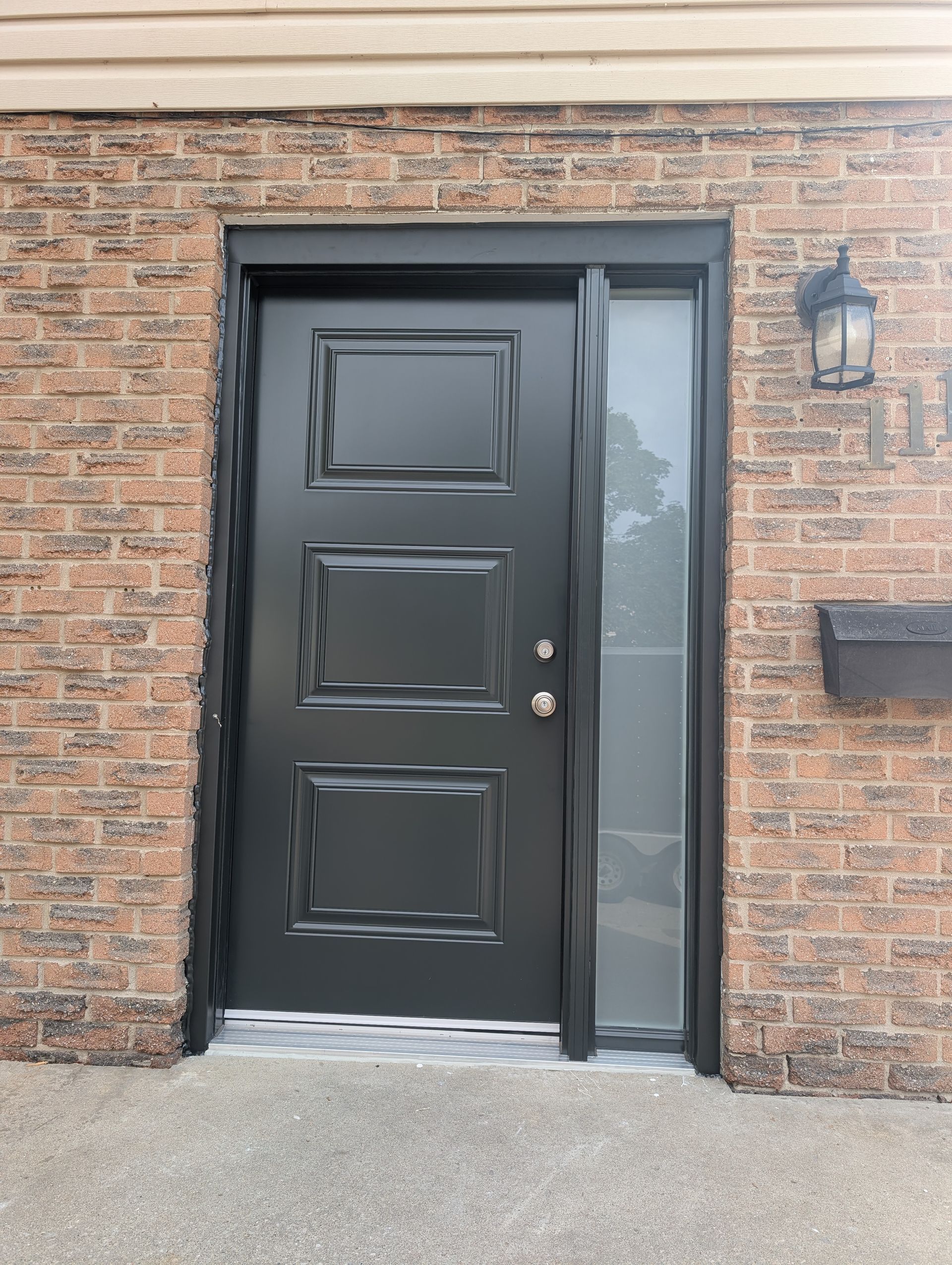 A black front door with three rectangular panels and a vertical sidelight set into a brown brick wall.