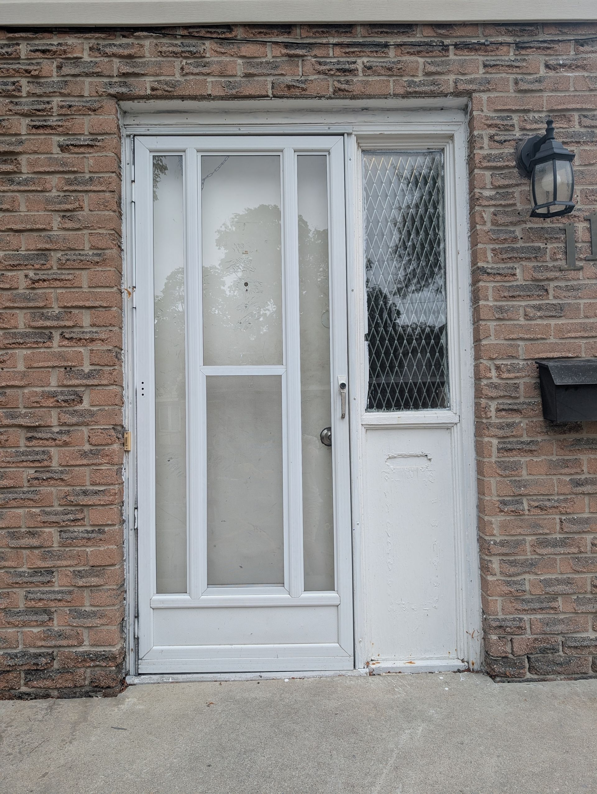 A white storm door with glass panels next to a sidelight with textured glass, set in a brown brick exterior wall.