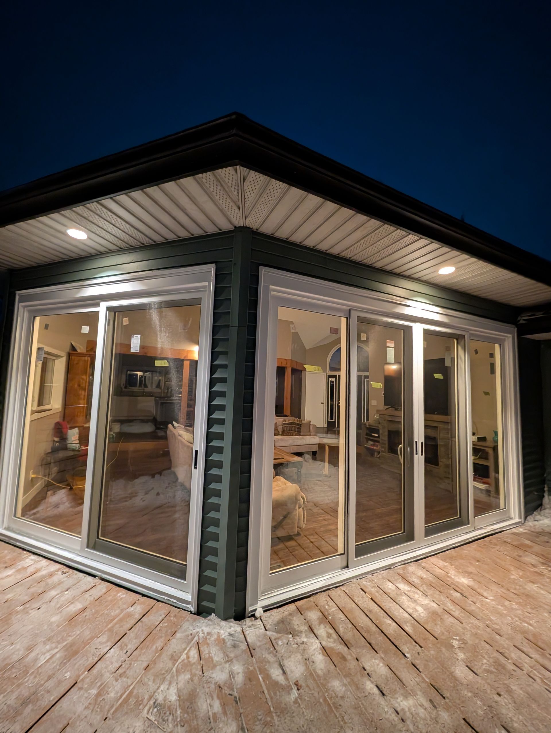 A corner view of a dark-sided house exterior at night, featuring large sliding glass doors leading to an illuminated deck.