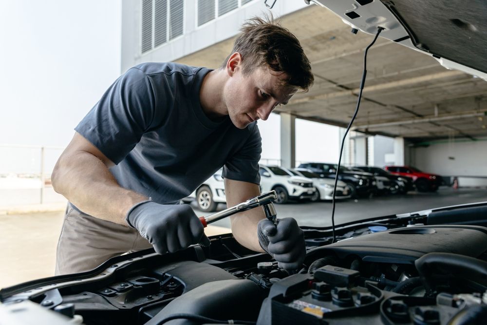 Mechanic with gloves working on a car engine in a parking garage. He is using a wrench, focused on the task.