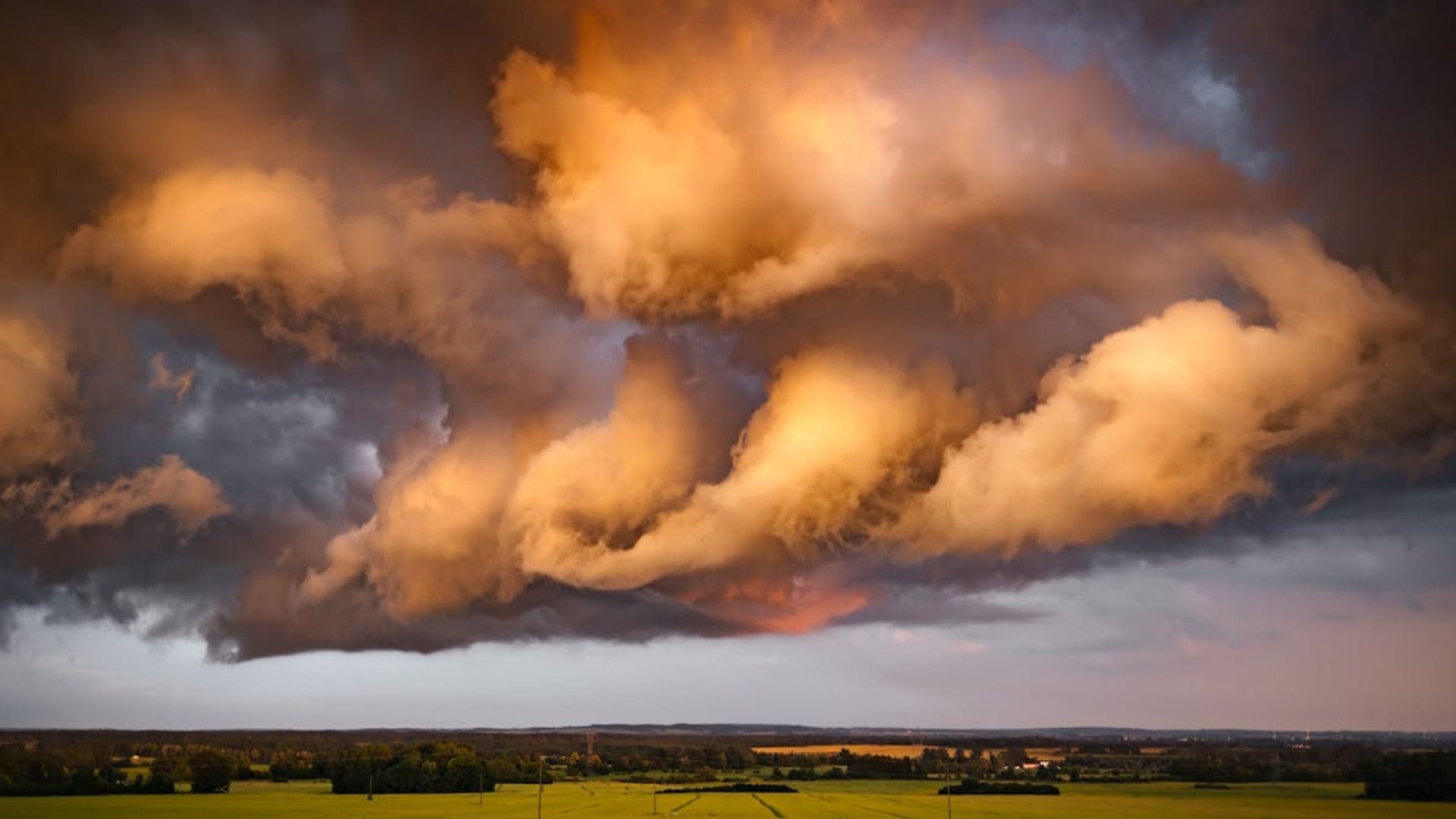 Nubes de tormenta dramáticas y brillantes sobre un campo verde y una línea de árboles distante, iluminadas por una luz cálida.