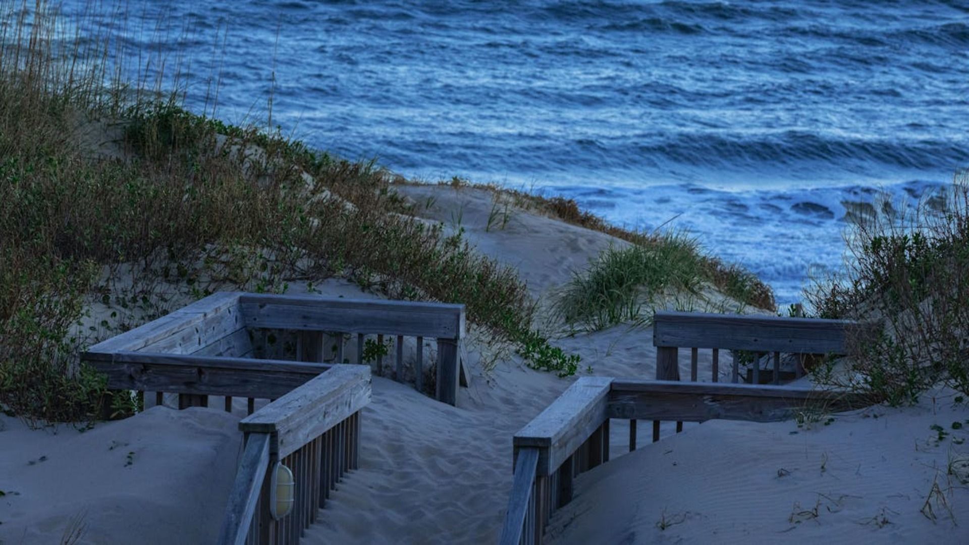 Camino de madera que conduce a una playa de arena y al océano bajo un cielo azul.