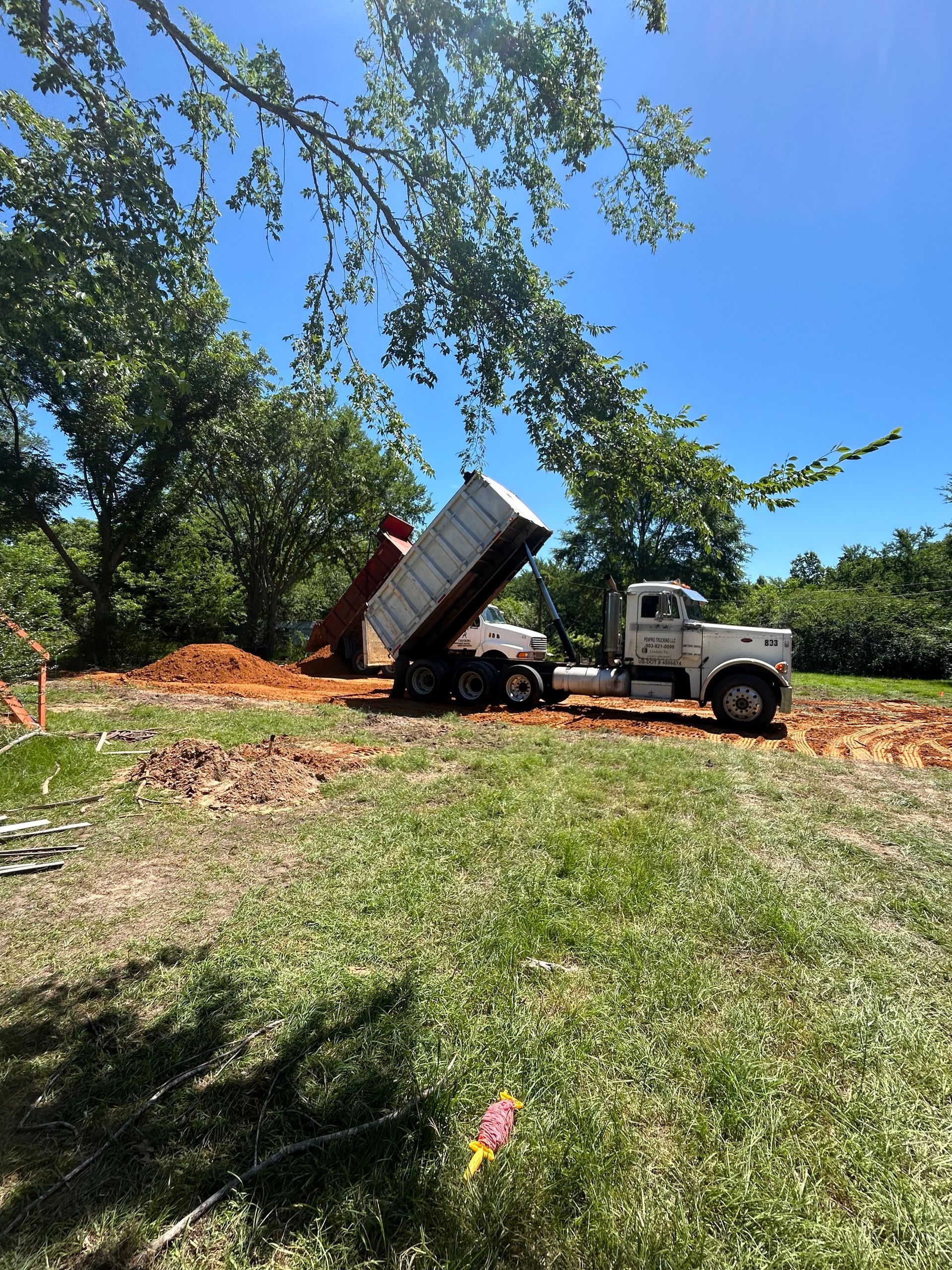 A dump truck dumping dirt onto a brown mound in a grassy area, with trees in the background under a blue sky.