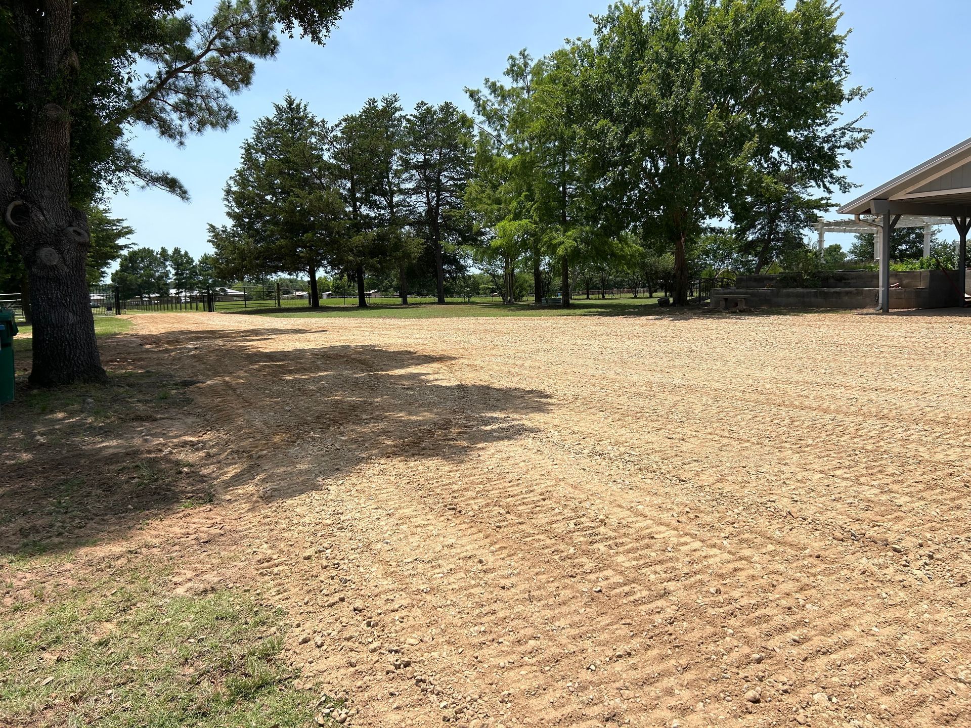Gravel driveway with tree shadows, a grassy lawn with trees in the background, and a building on the right.