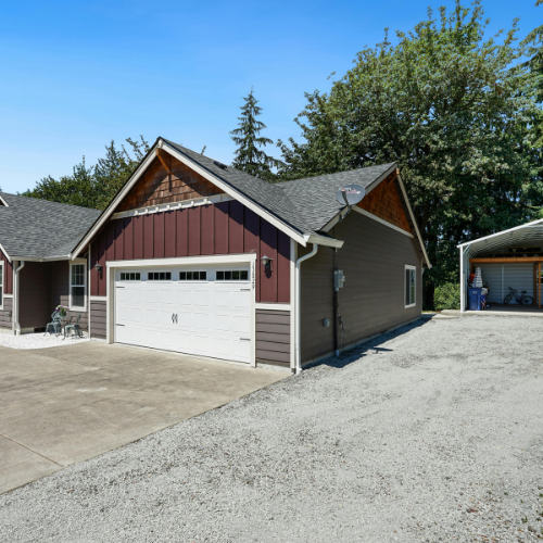 Garage with a gravel driveway and carport on a sunny day. Brown and gray siding.