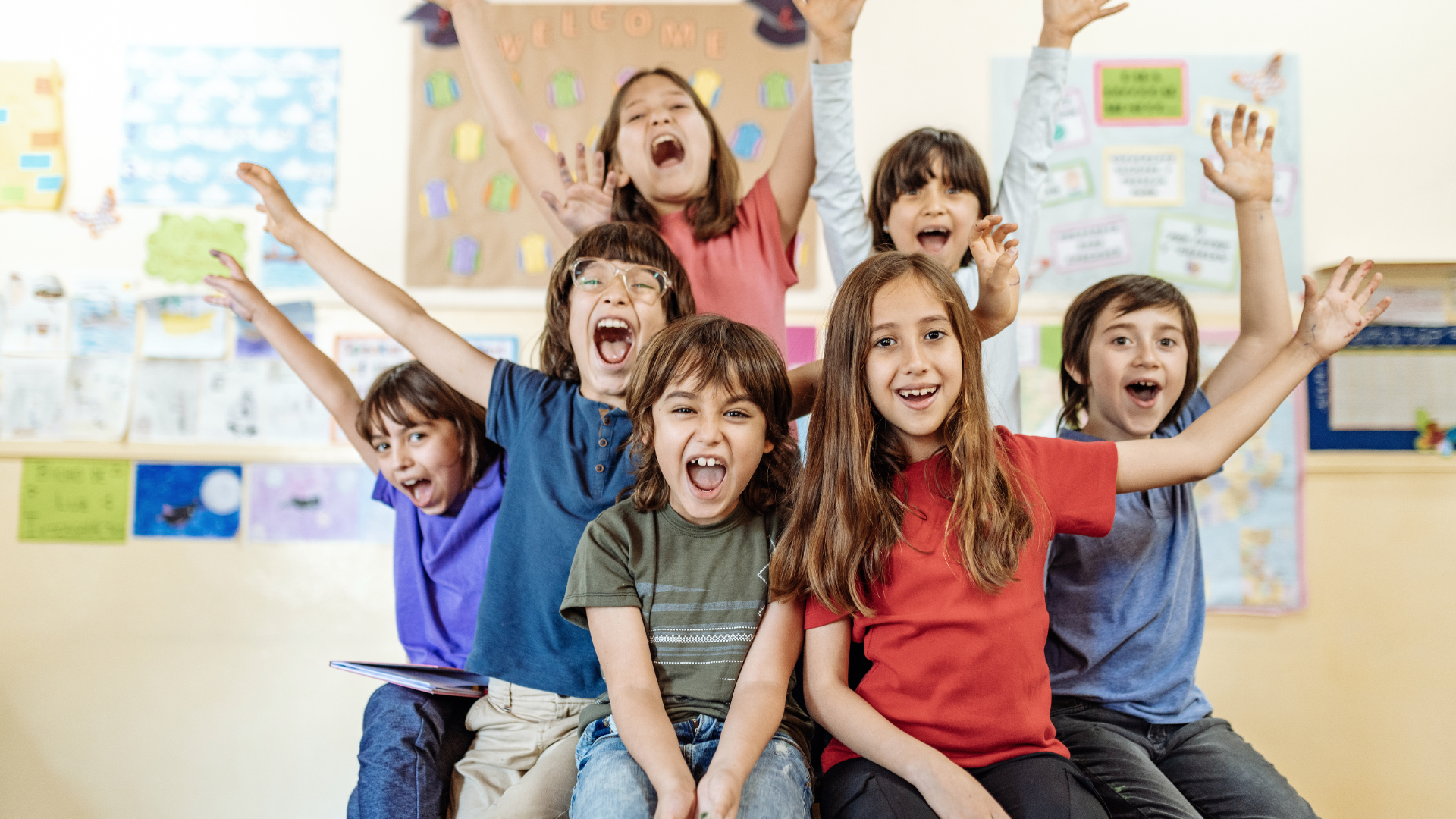 A group of children are sitting on the floor in a classroom with their arms in the air.