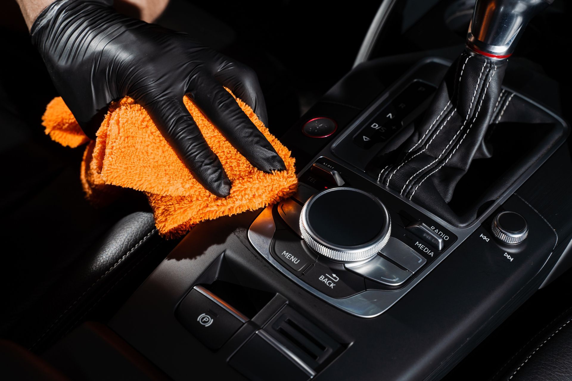 Black-gloved hand wiping a car’s center console with an orange microfiber cloth