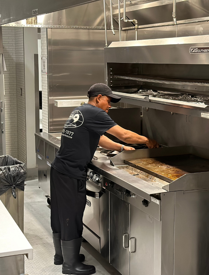 A man is standing in a kitchen scraping and cleaning a grill in preparation for sanitization. 