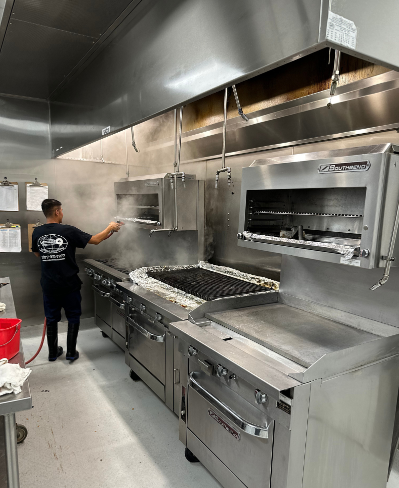 A man in a black Gemilang Solutions shirt is sanitizing a stove and ventilation system in a kitchen.