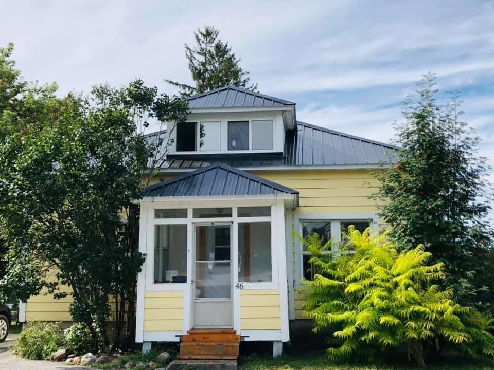 Une maison jaune avec un toit noir est entourée d'arbres et de buissons.
