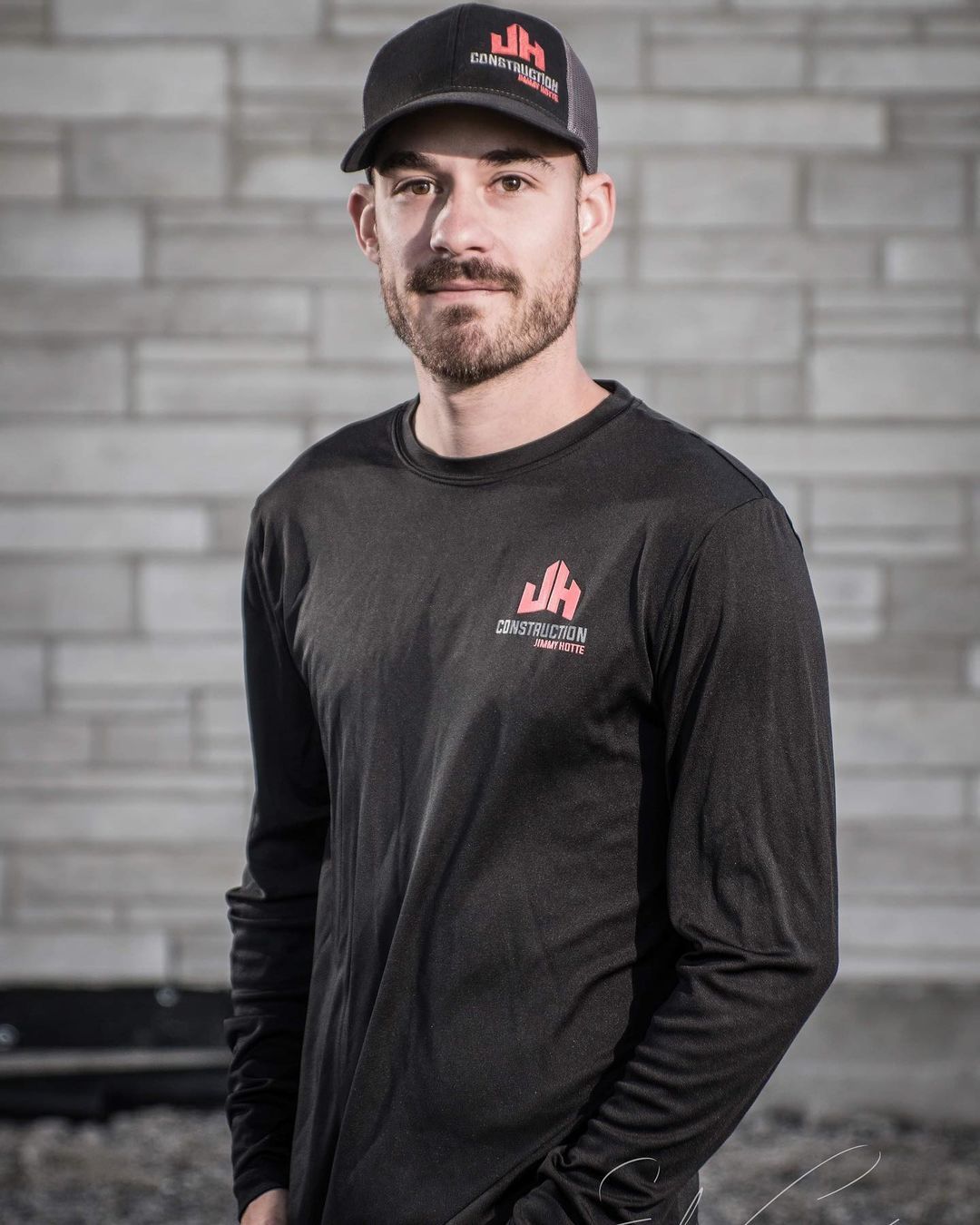 Un homme portant un chapeau et une chemise à manches longues se tient devant un mur de briques.