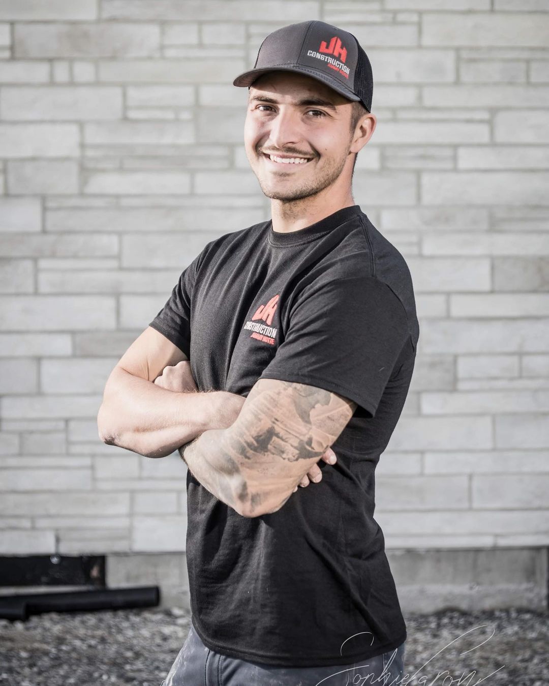 Un homme portant un chapeau et une chemise noire se tient debout, les bras croisés devant un mur de briques.