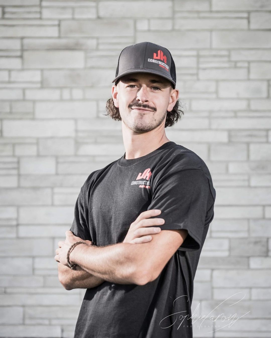 Un homme portant un chapeau et une chemise noire se tient debout, les bras croisés devant un mur de briques.