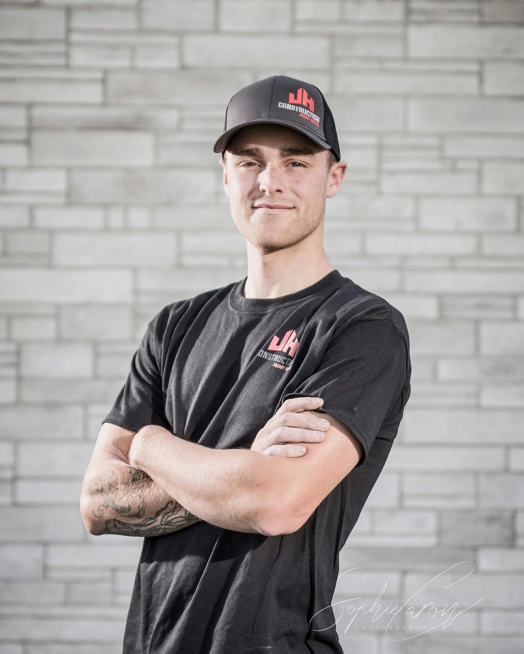 Un homme portant un chapeau et une chemise noire se tient debout, les bras croisés devant un mur de briques.