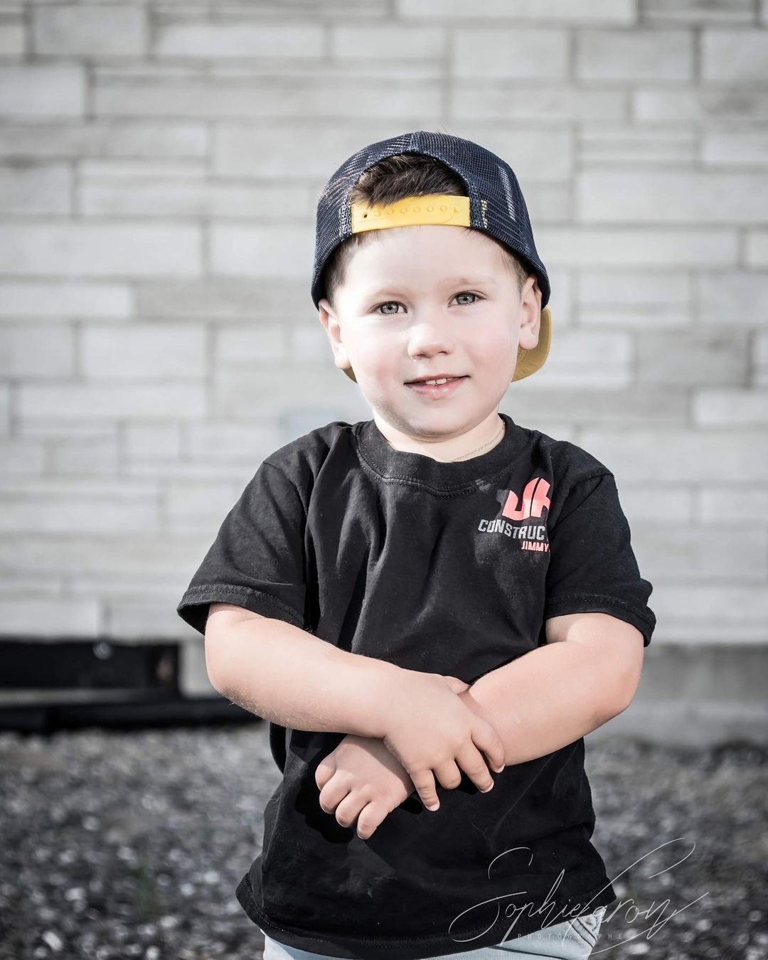 Un jeune garçon portant un chapeau et une chemise noire se tient devant un mur de briques.