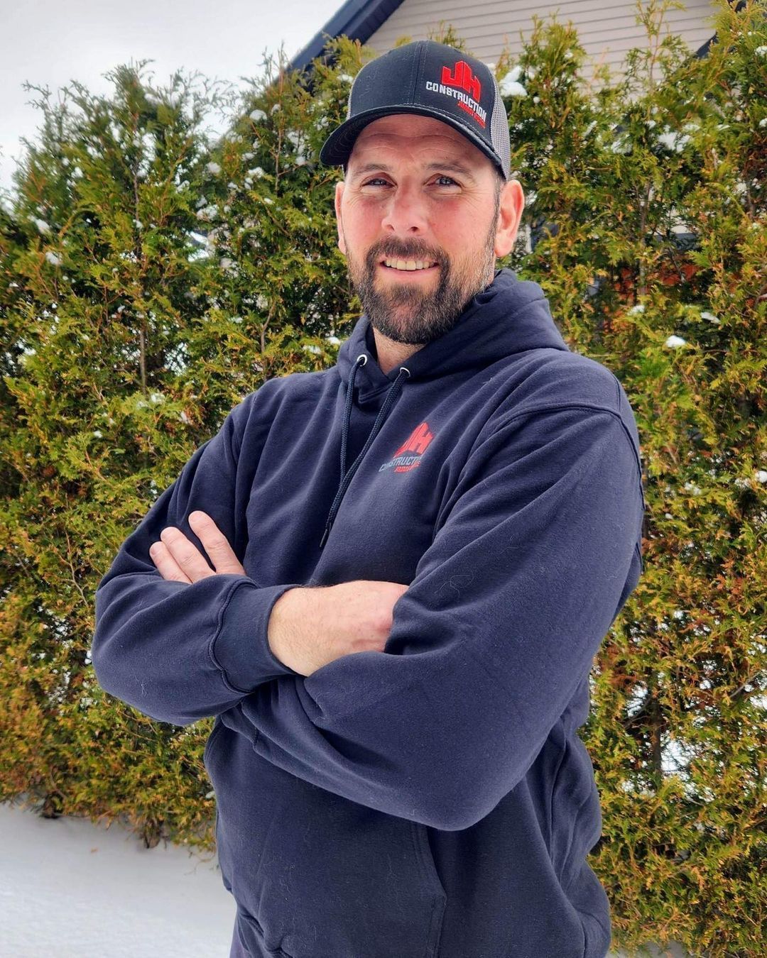 Un homme avec une barbe et un chapeau se tient debout, les bras croisés, devant un buisson.