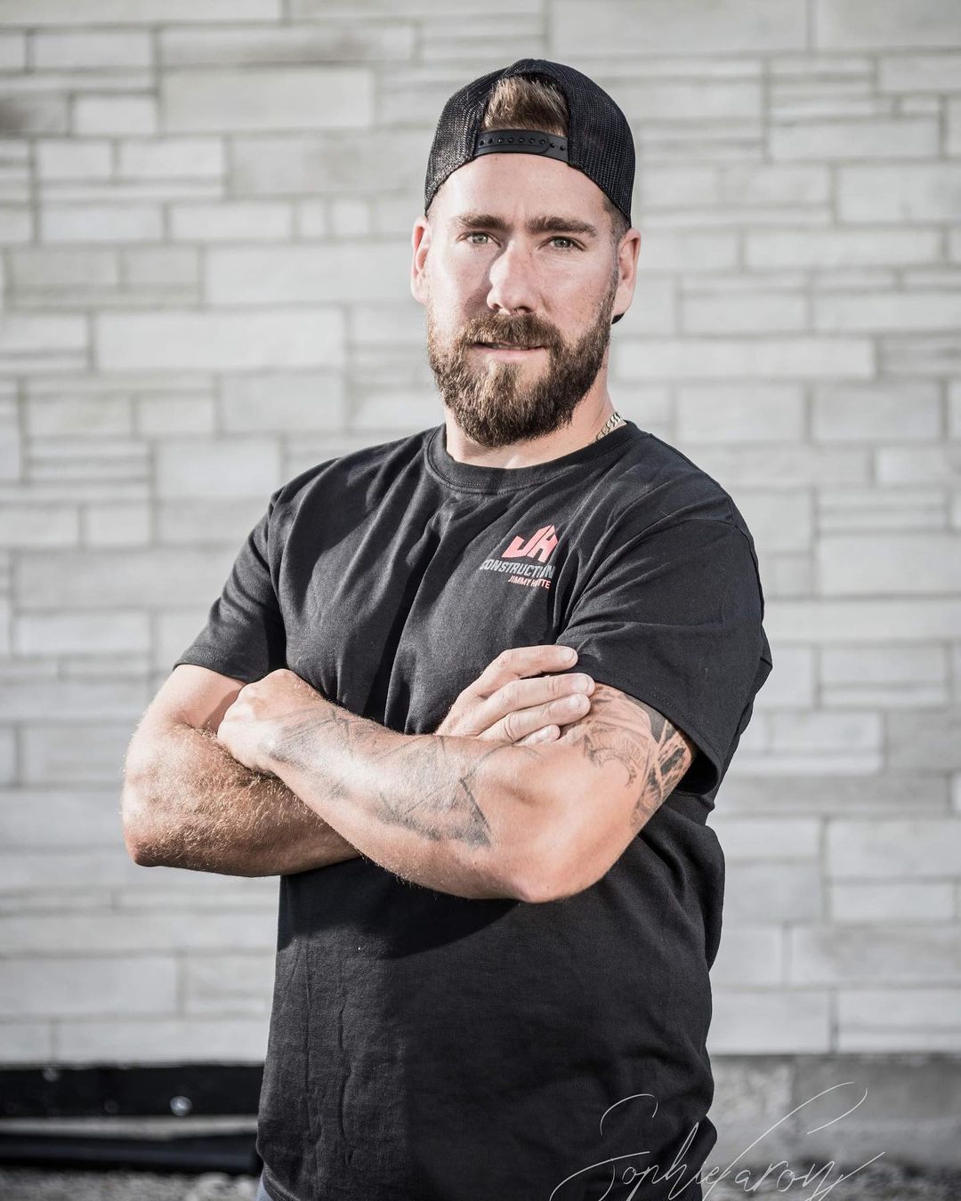Un homme avec une barbe et des tatouages ​​se tient debout, les bras croisés devant un mur de briques.