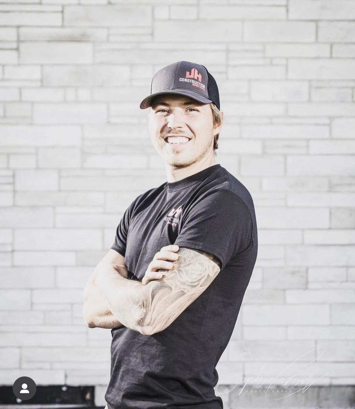 Un homme portant un chapeau et une chemise noire se tient debout, les bras croisés devant un mur de briques.