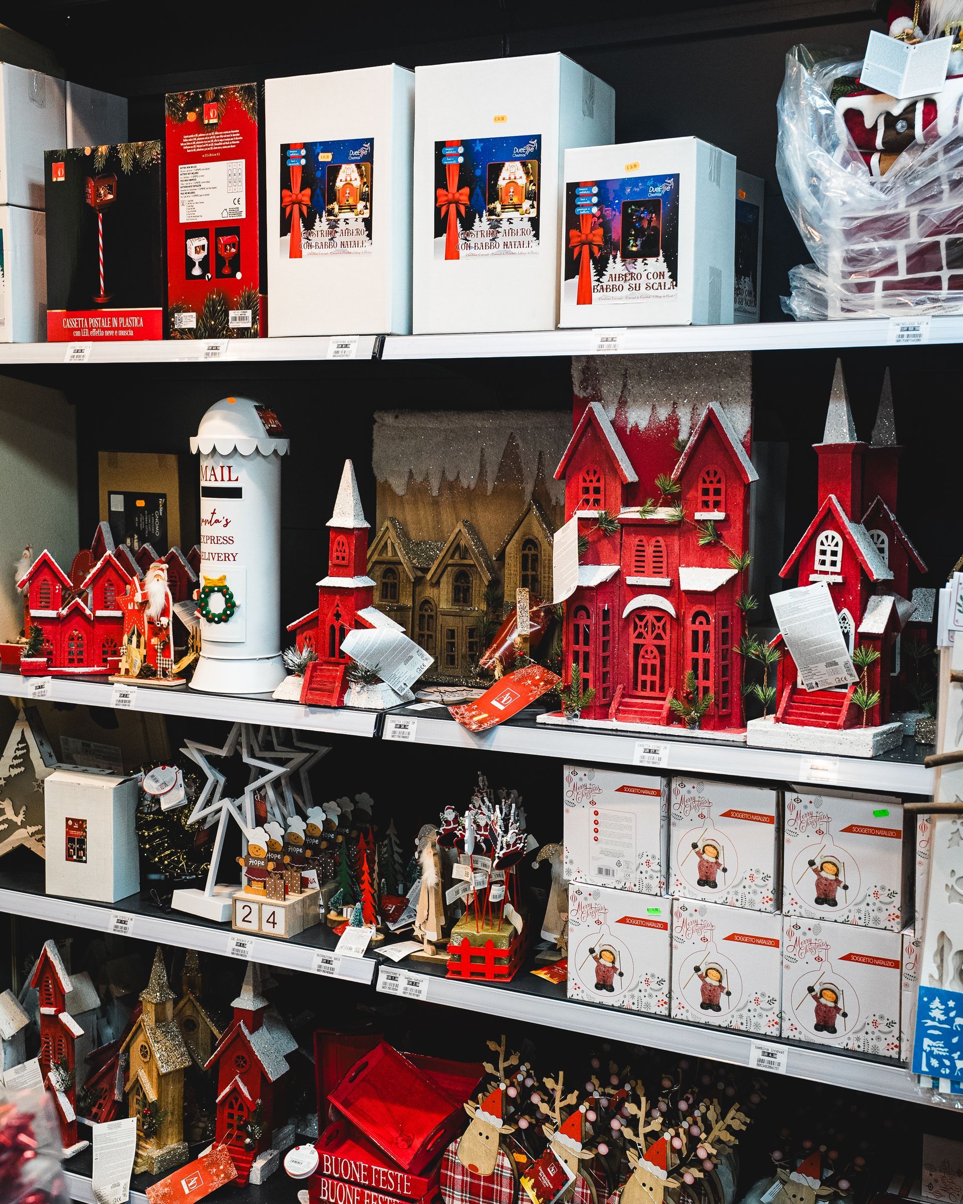 Scaffali pieni di decorazioni natalizie rosse e bianche, tra cui casette, statuette e scatole.