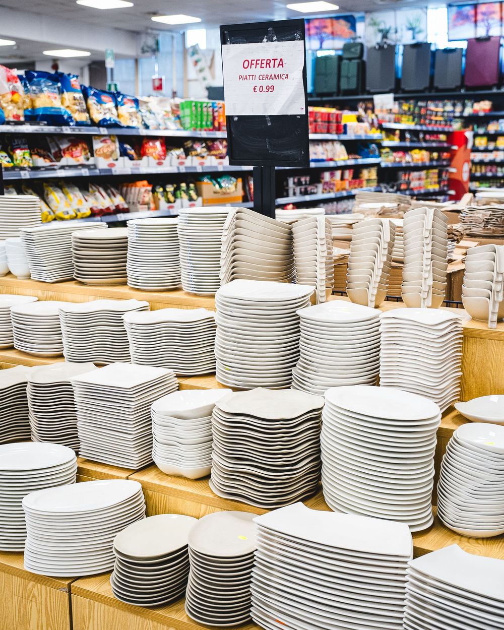 Pile di piatti bianchi esposti in un negozio, con scaffali di cibo confezionato sullo sfondo.