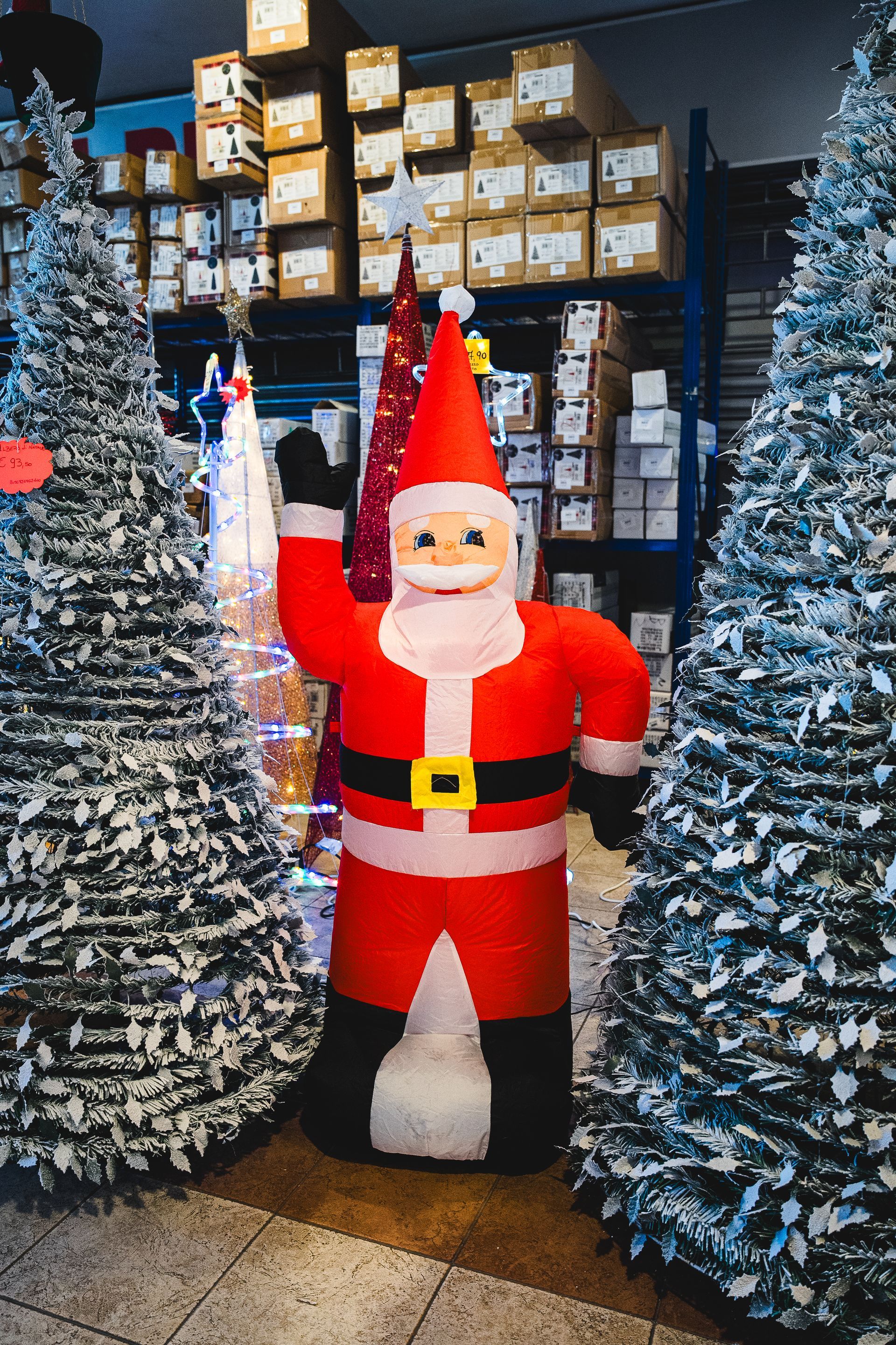 Babbo Natale gonfiabile che saluta tra due alberi di Natale innevati, con scatole sugli scaffali sullo sfondo.