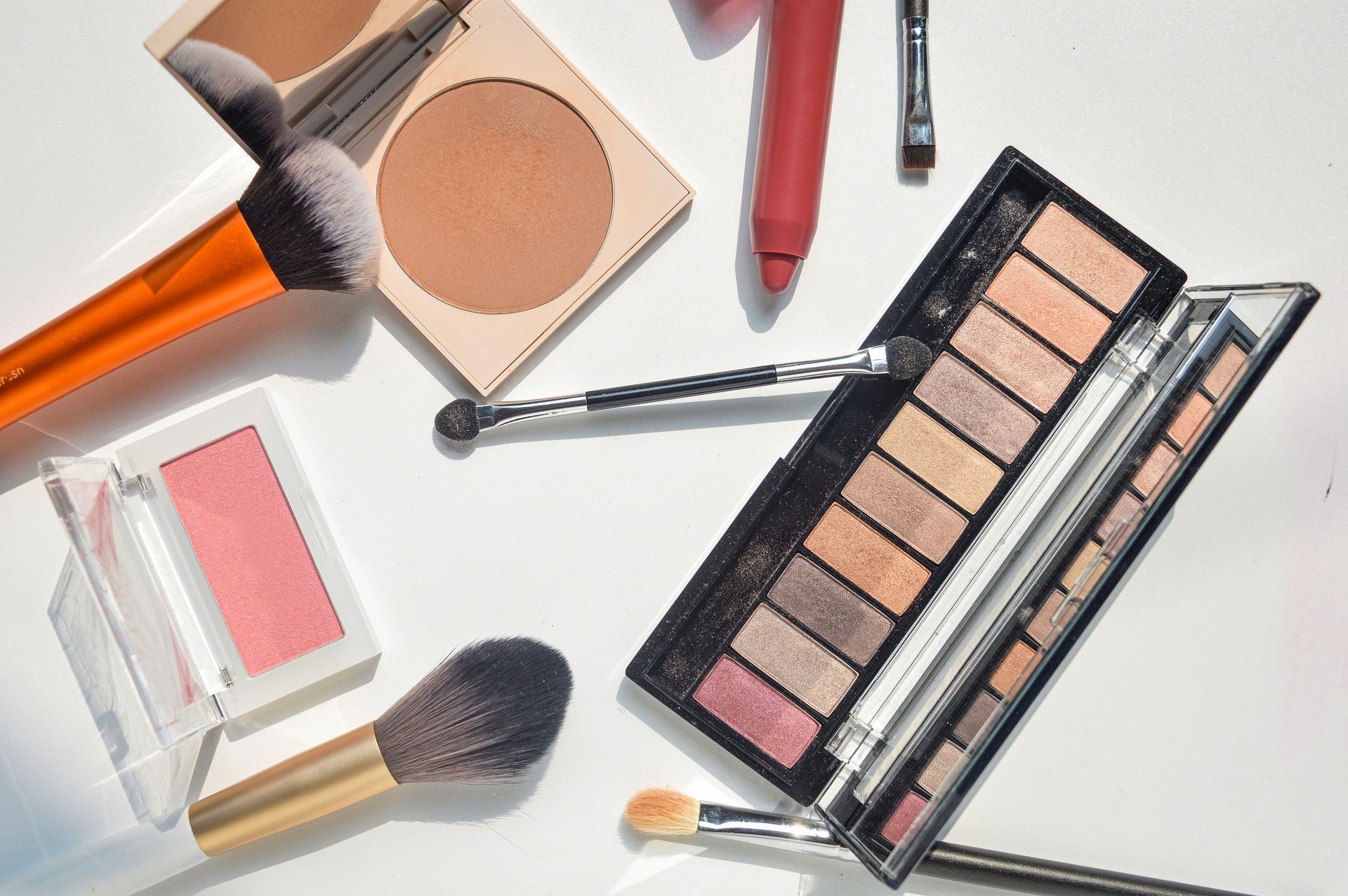 Prodotti per il trucco, tra cui palette di ombretti, fard, terra abbronzante, matita labbra e pennelli, su una superficie bianca.