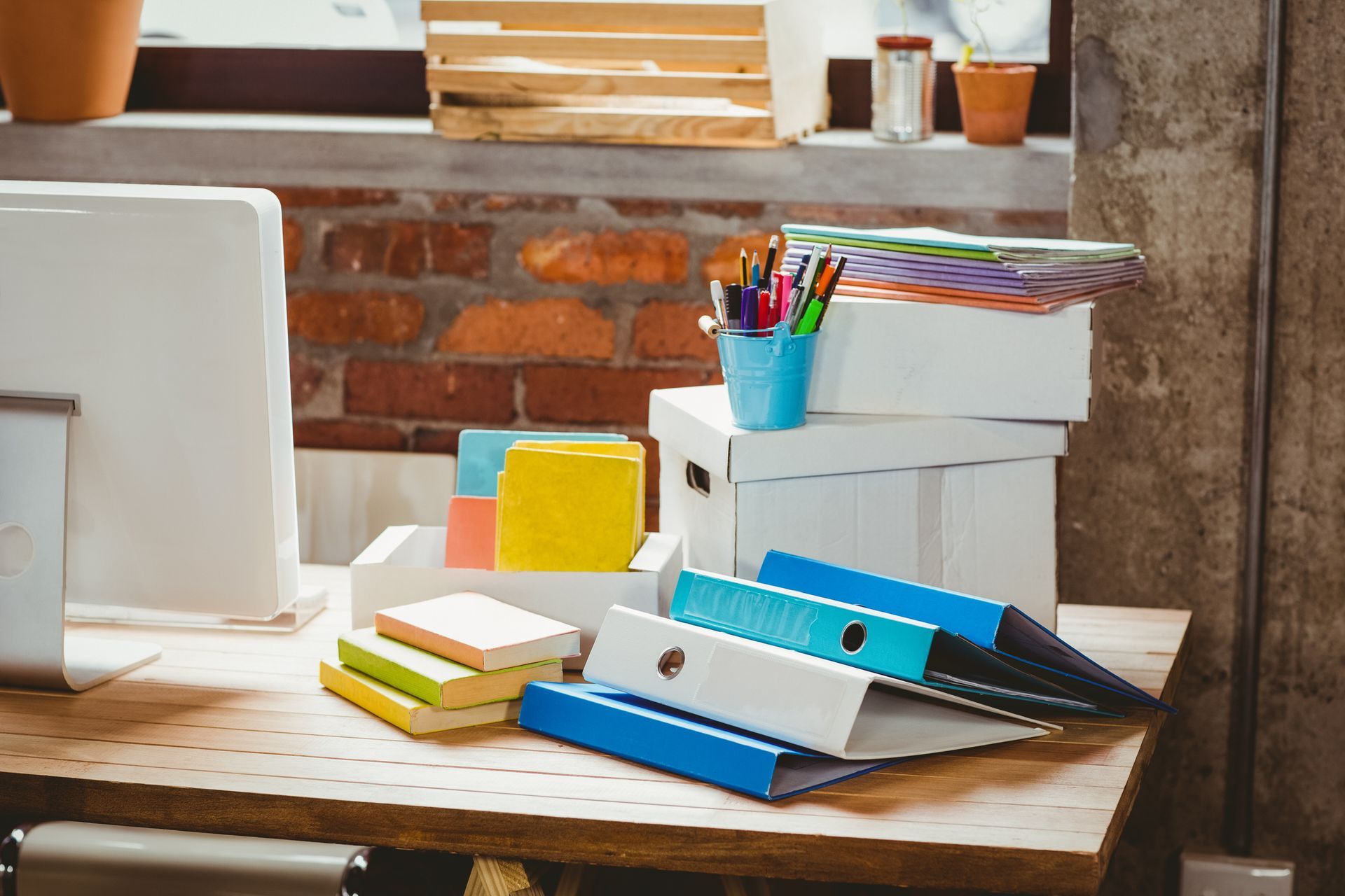Scrivania con materiale per ufficio: scatole impilate, raccoglitori, penne, carta. Muro di mattoni sullo sfondo.