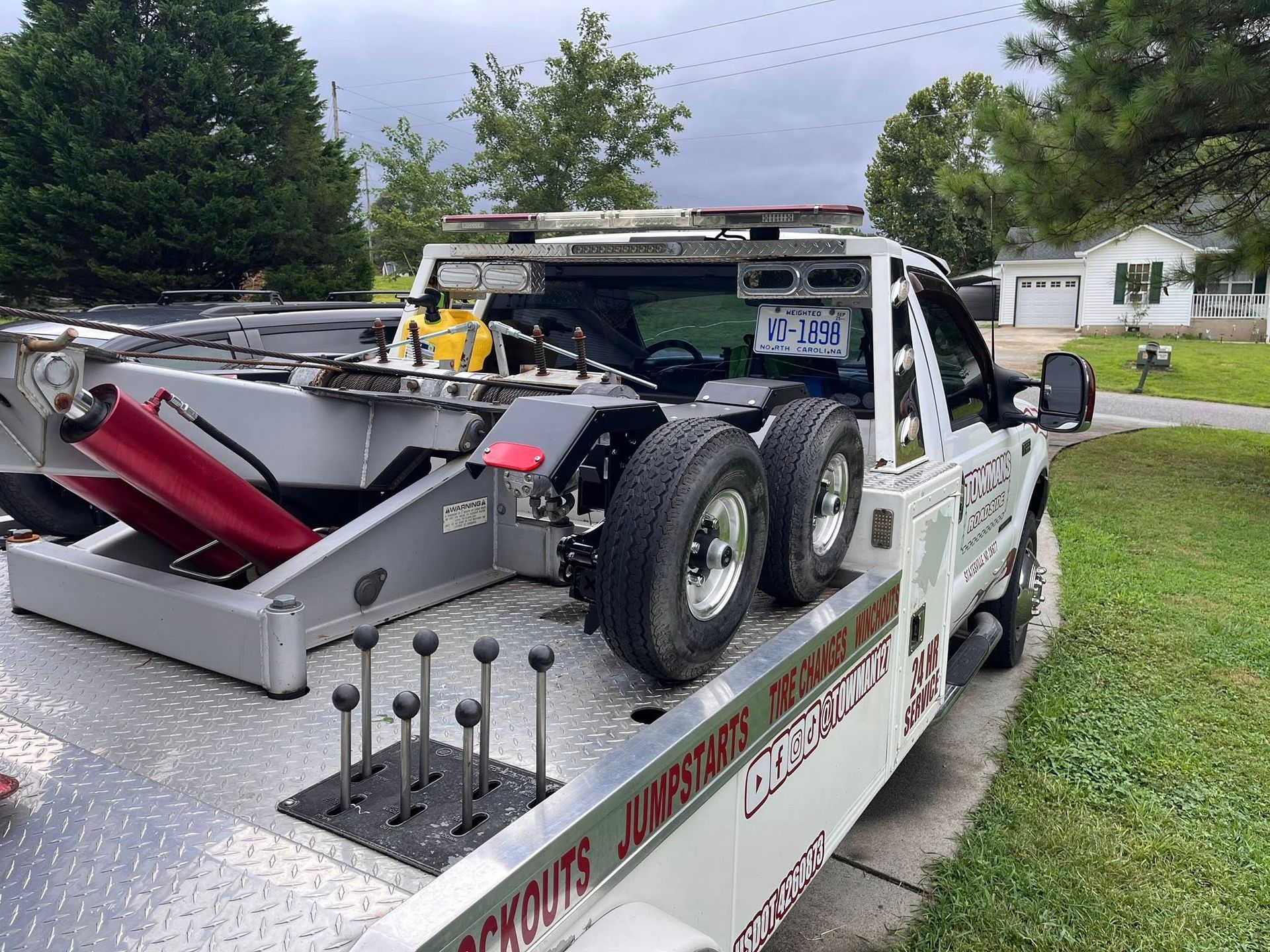 Tow truck on a residential street. White vehicle with tires and towing equipment. Green grass and houses in the background.
