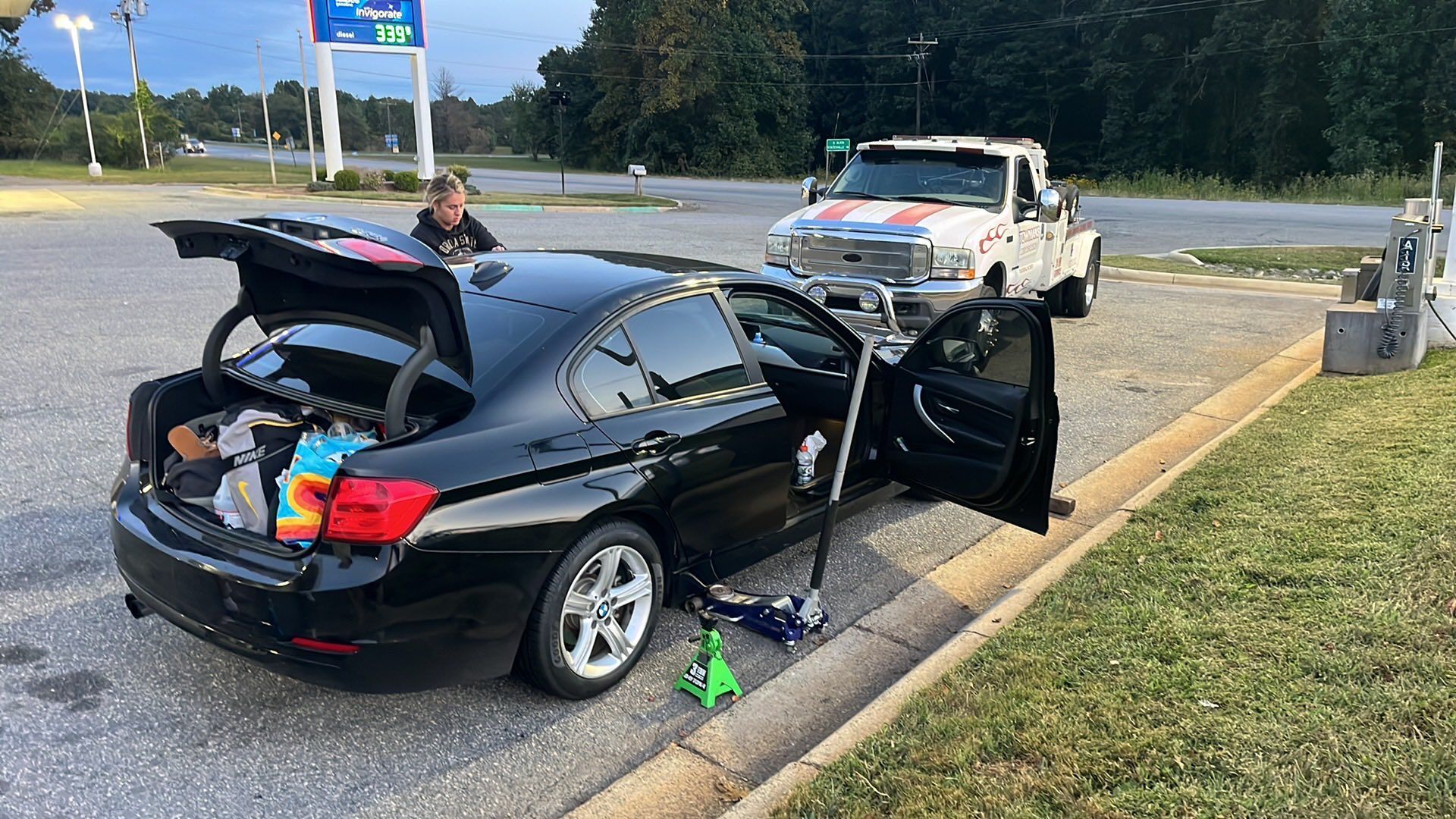 Black car with open trunk and door, jacked up on roadside near gas station, tow truck present.