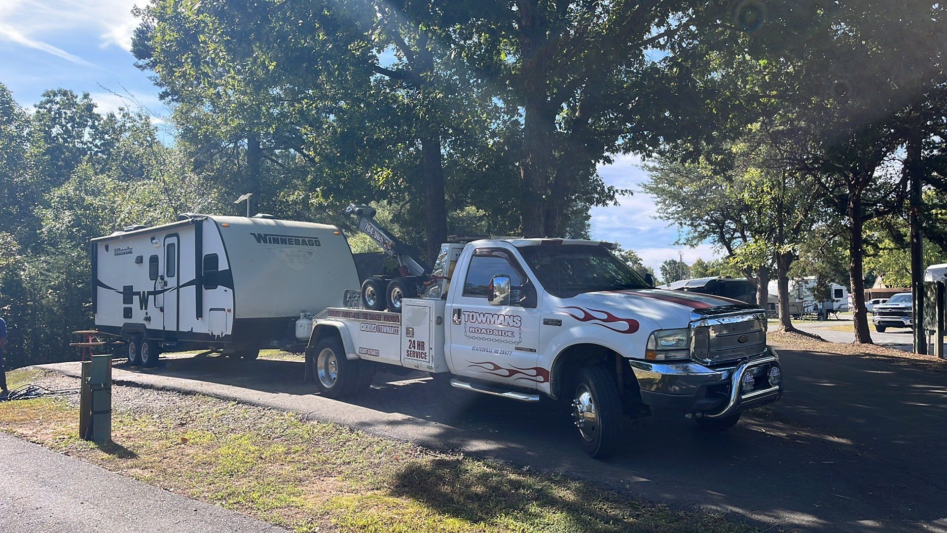 Tow truck with a camper trailer parked on a grassy campground under trees on a sunny day.