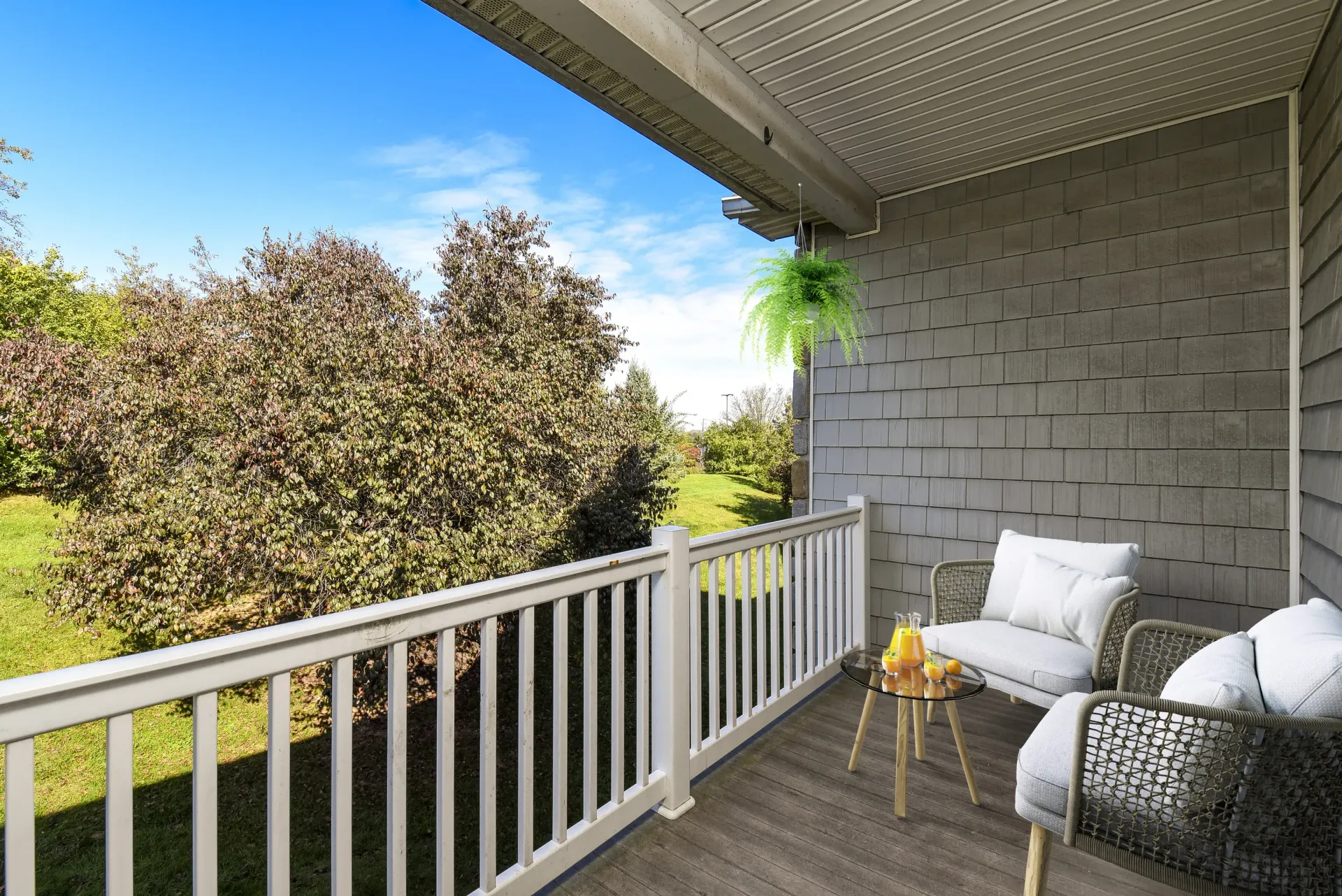 Balcony with cushioned wicker seating, a small round table, a hanging fern, and a view of a green lawn at Springhouse Townhomes in Allentown, PA.