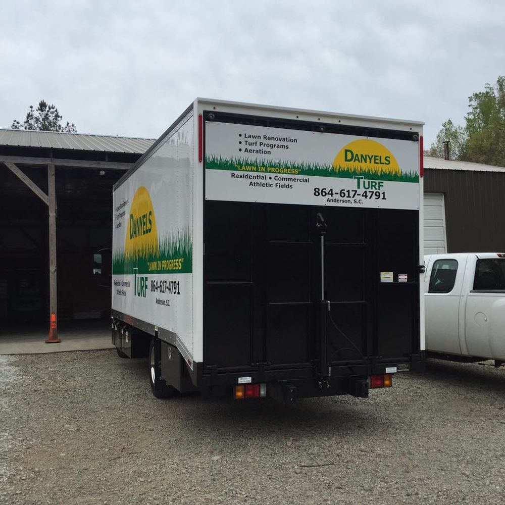 White Daniels Turf delivery truck with black back doors, parked in a gravel lot.