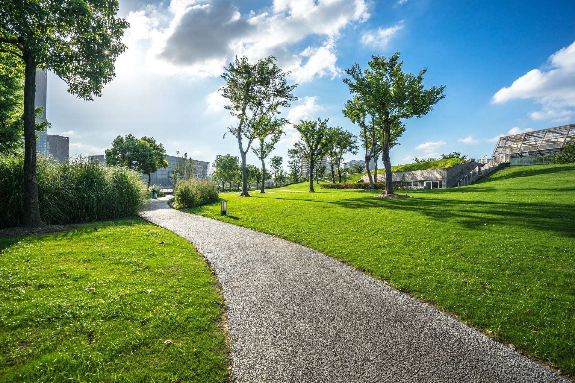 Stone path winds through a lush green park with trees, under a blue sky with clouds.