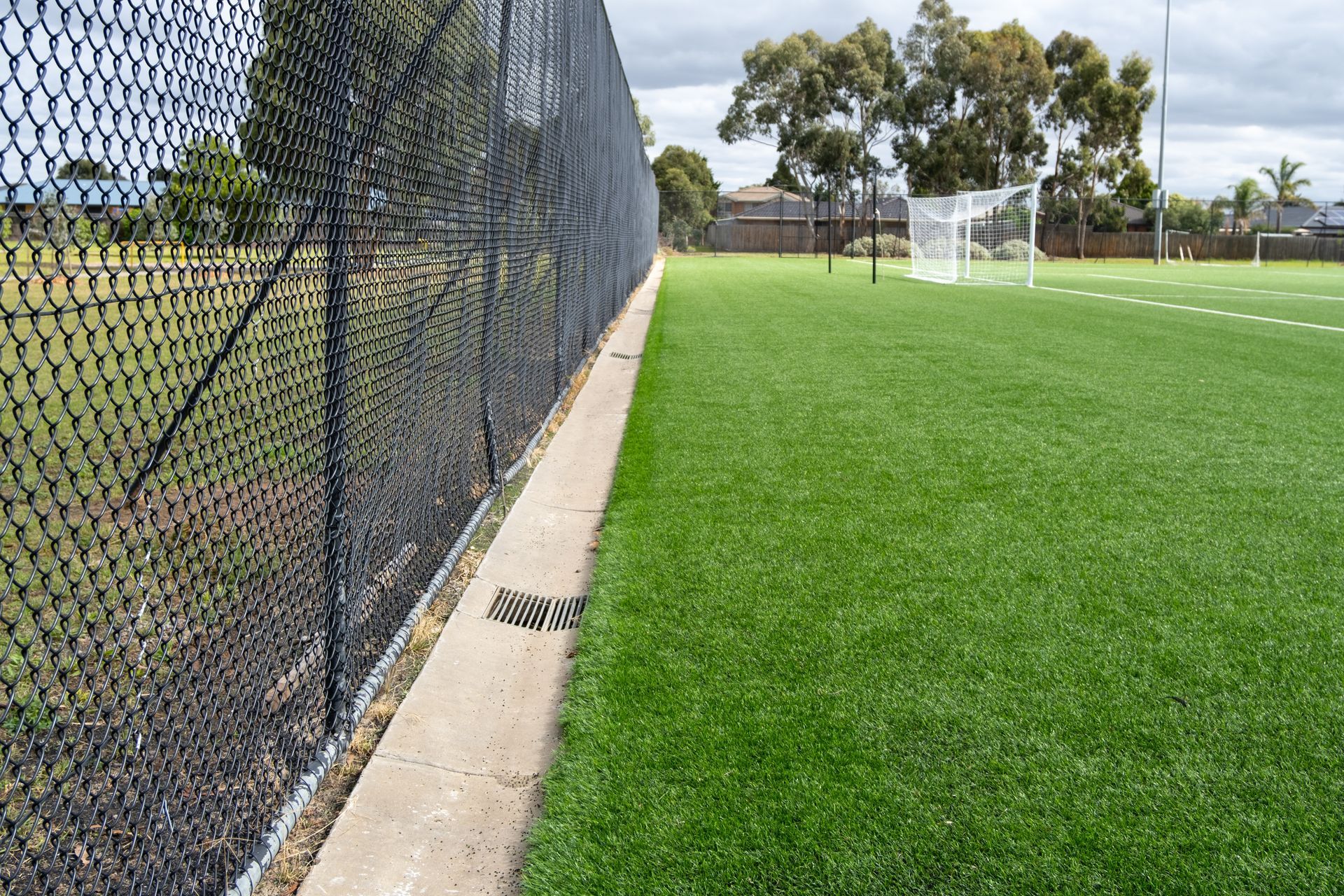 Black chain-link fence bordering a green artificial turf soccer field under a cloudy sky.