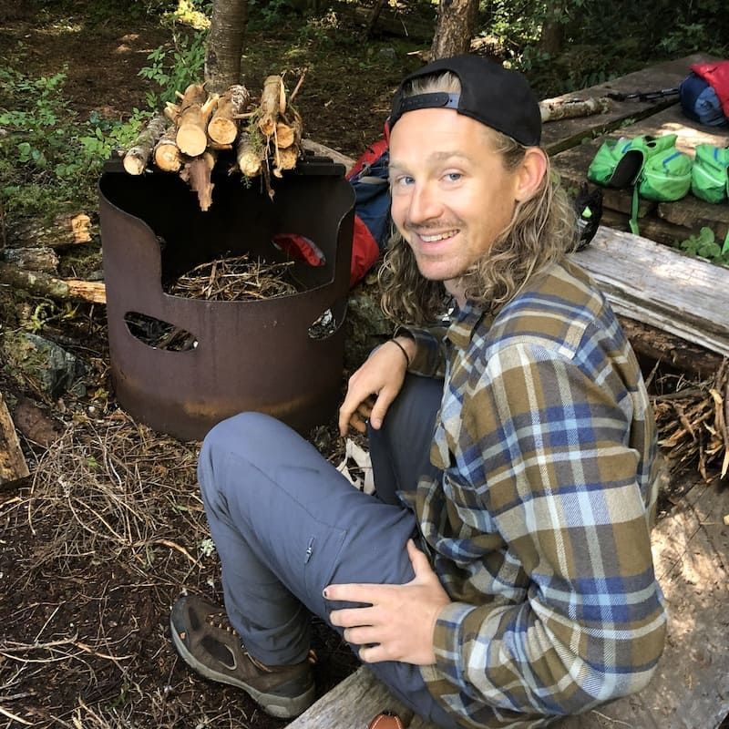 A man in a plaid shirt is sitting on a bench in front of a pile of logs.