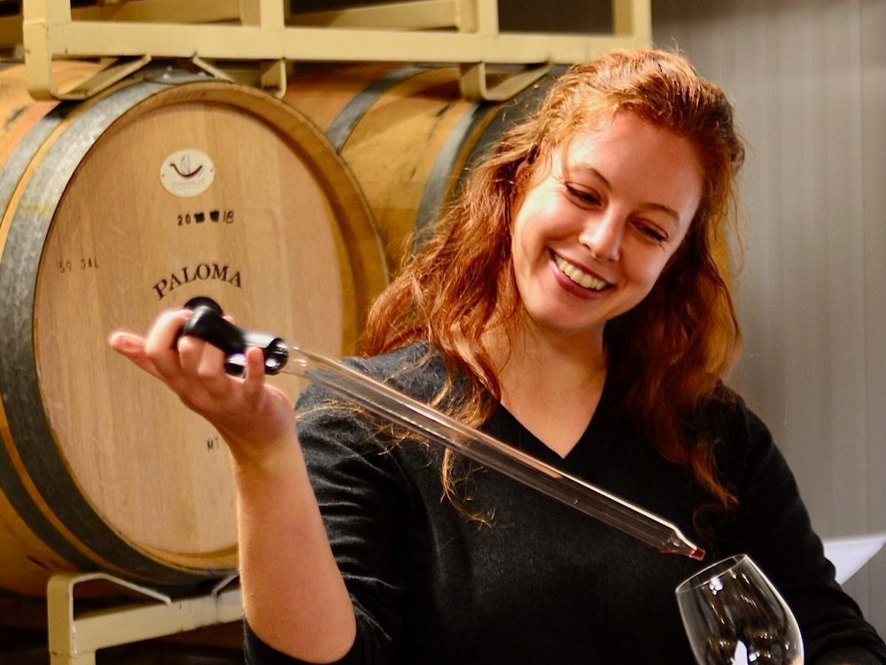 A woman is pouring wine into a glass in front of barrels that say paloma