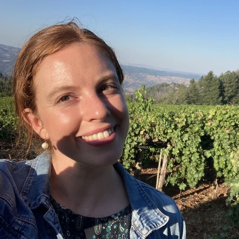A woman in a denim jacket is smiling in front of a vineyard
