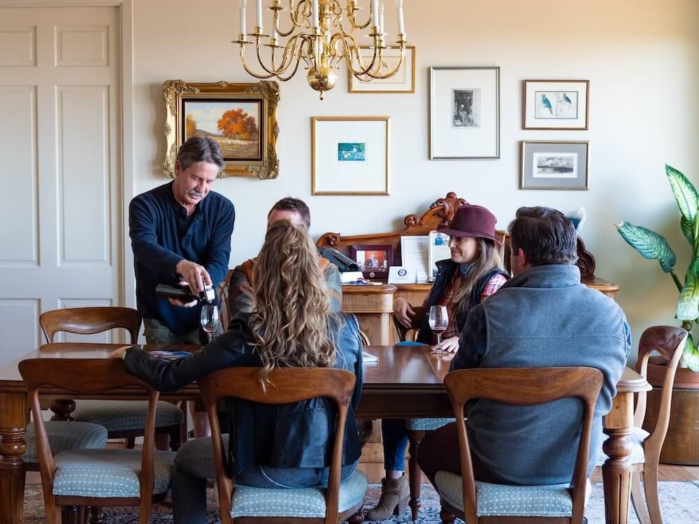 A man is pouring wine into a glass for a group of people sitting at a table.