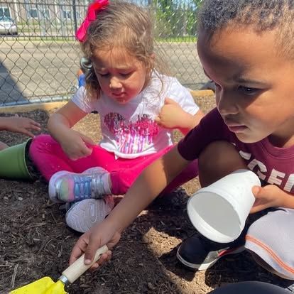 A boy and a girl are playing in the dirt with a shovel and cups.