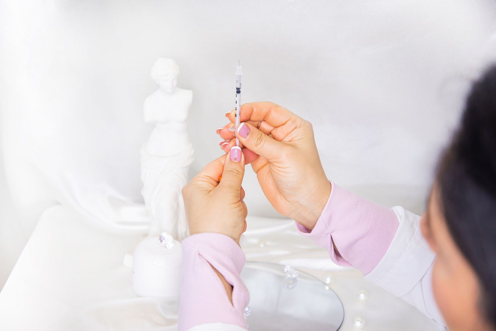 Person holding a syringe, preparing for injection, with a white statue and draped fabric in the background.