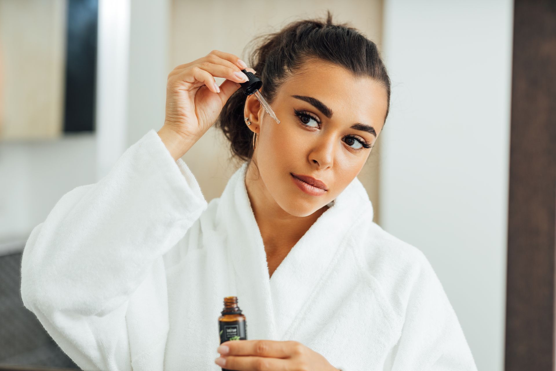 A woman in a bathrobe is applying a serum to her face.