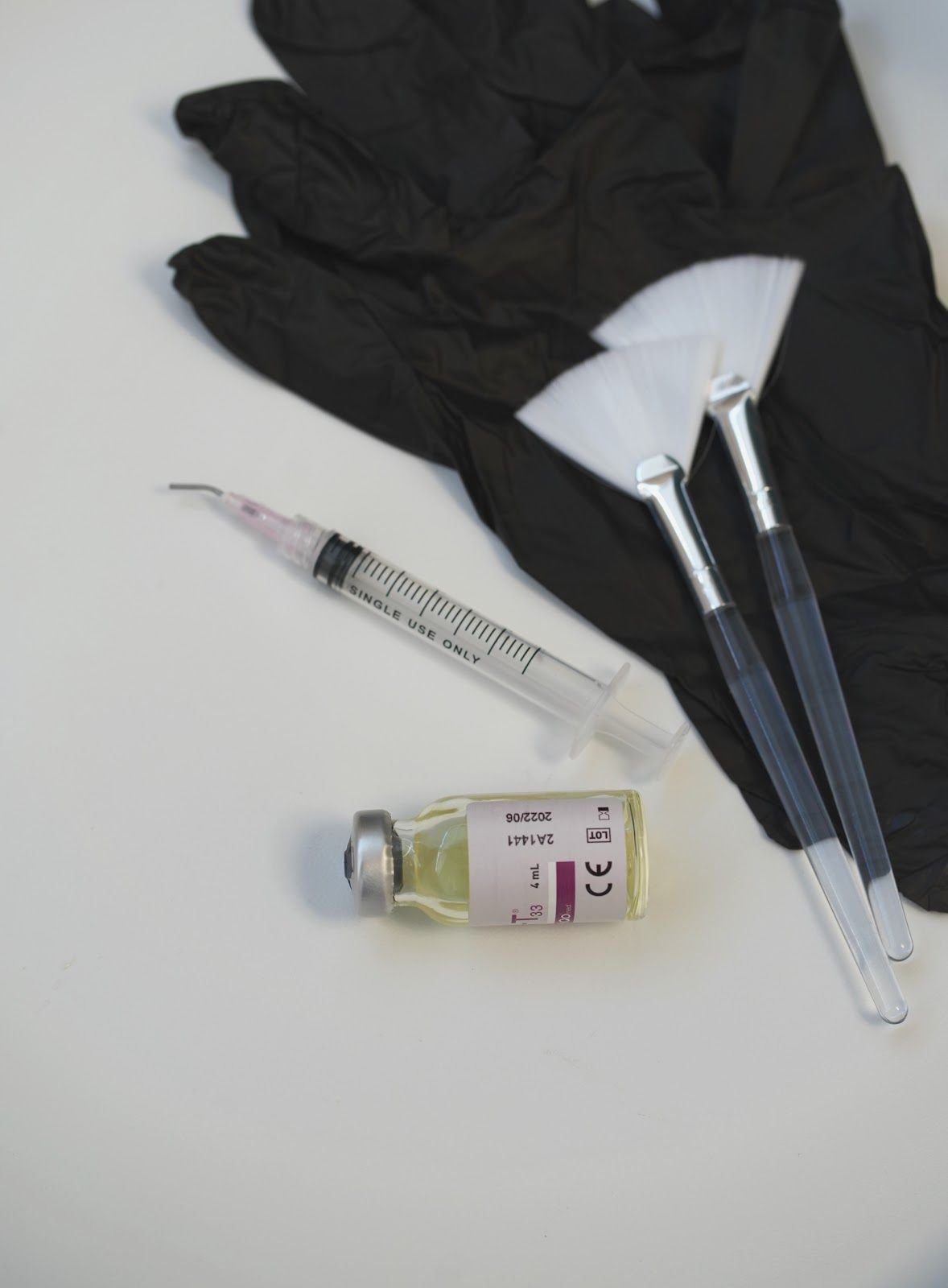 Black gloves, syringe, small bottle, and two fan-shaped brushes on a white surface, possibly for cosmetic procedures.
