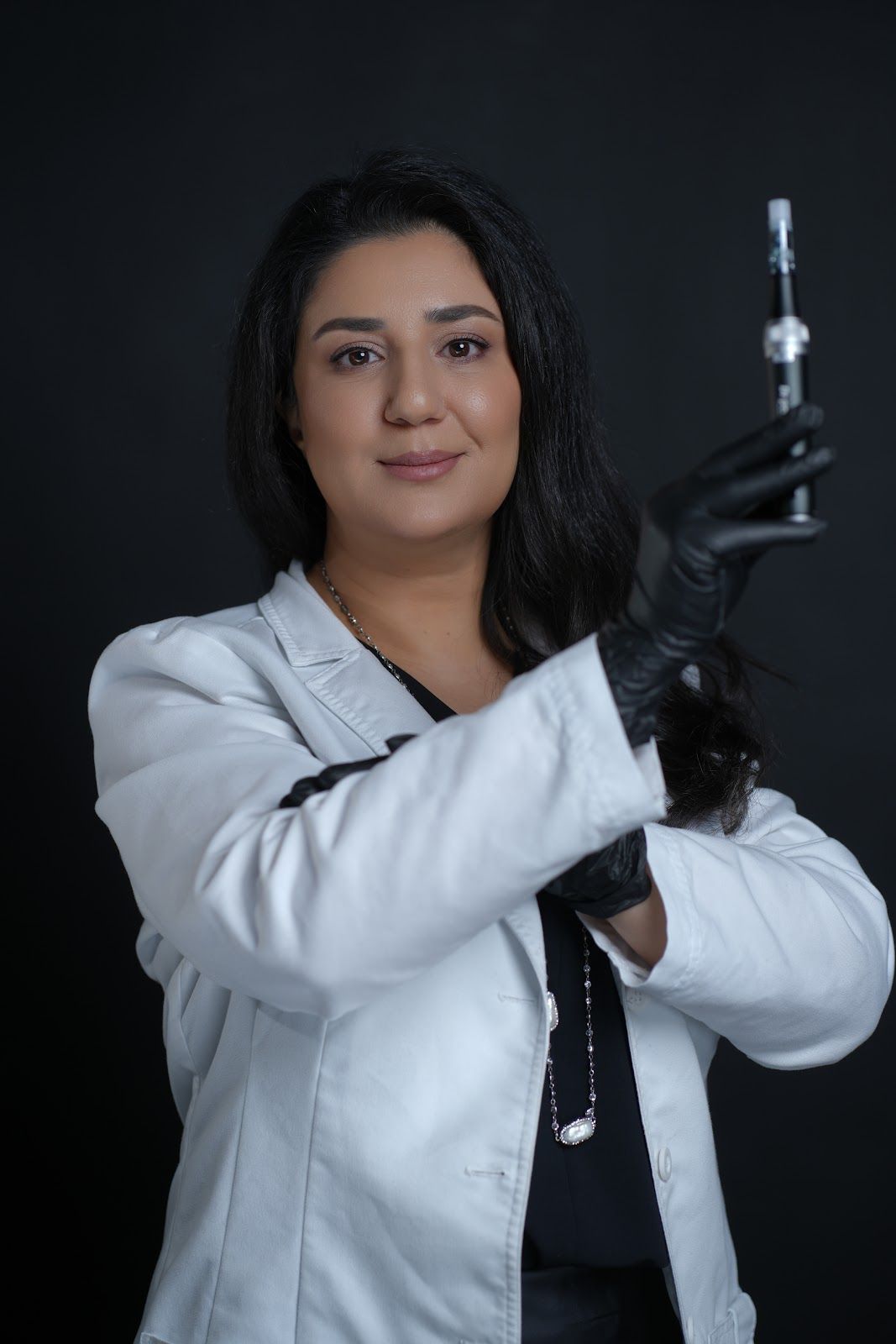 Woman in lab coat and gloves holding syringe, dark background.