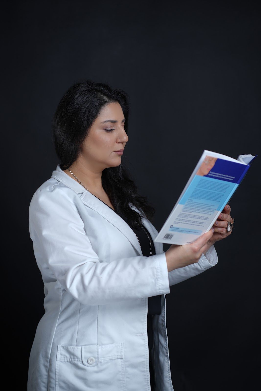 Woman in white lab coat reads a book against a black background.