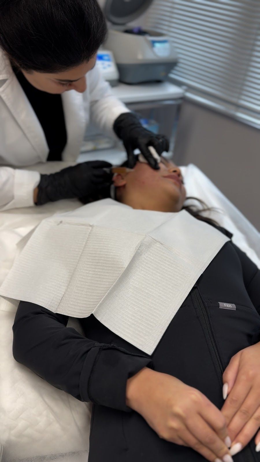 A medical professional in a lab coat performs a facial injection on a patient lying down. Both wear black gloves; the setting is a medical office.