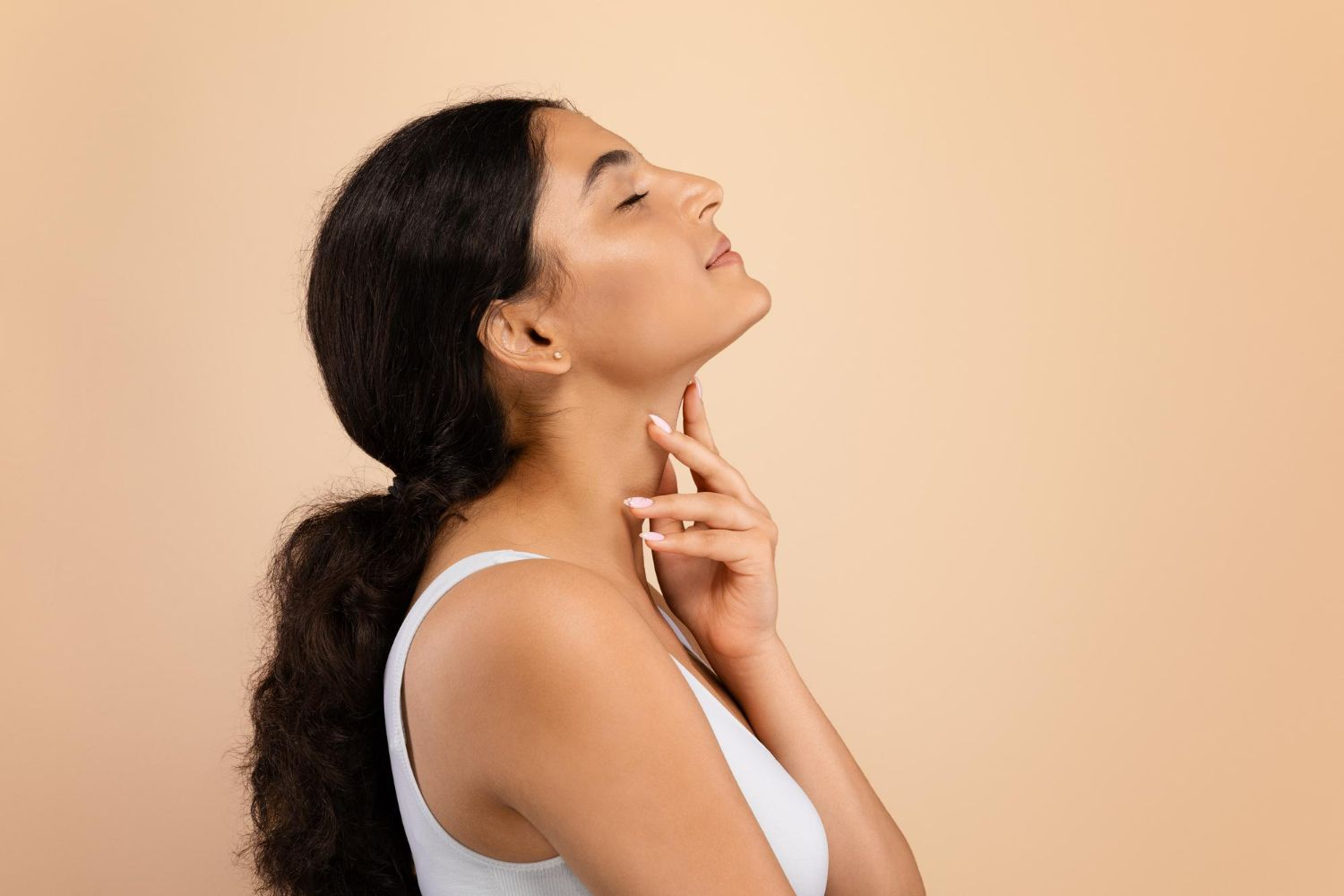 Woman with dark hair, eyes closed, hand on her neck, against a beige background.