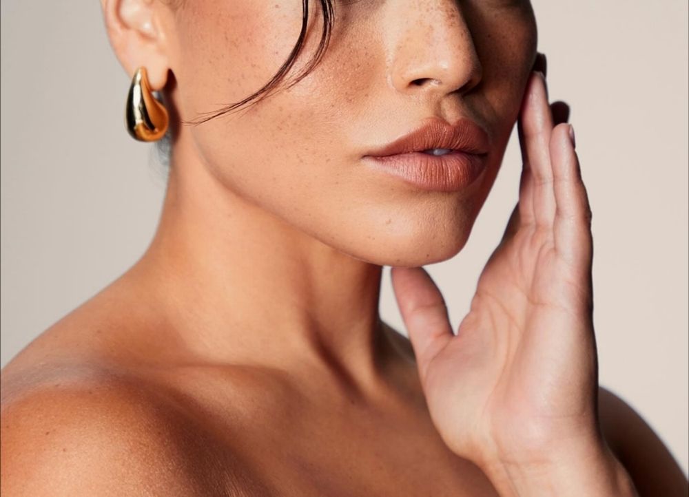 Close-up of a person with tanned skin, gold hoop earrings, and hand gently touching their face, against a neutral background.