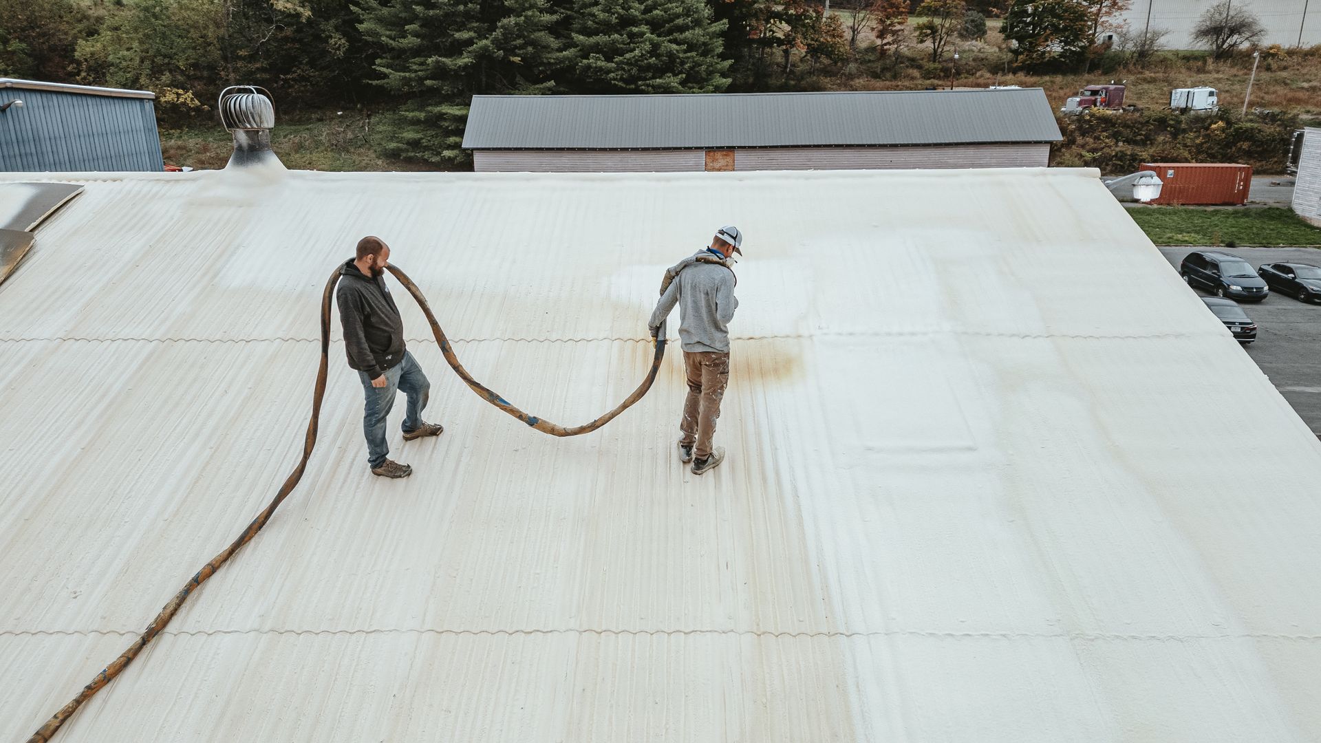 Two people applying a brown sealant to a white roof with a hose-like applicator outdoors.