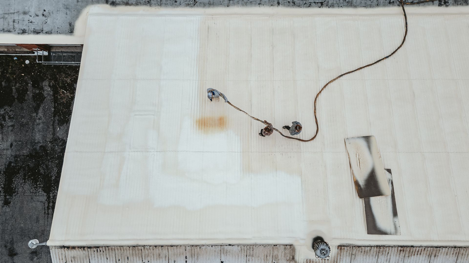 Workers on a large flat roof, applying a white coating with equipment and tools.